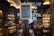 A cozy and rustic store interior with wooden shelves filled with various bottles and jars on display. A chalkboard sign stands in the center, listing items like Ibérico charcuterie and wines. Warm lighting enhances the inviting atmosphere, and a black tiled wall features illuminated text in the background.
