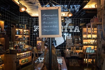 A cozy and rustic store interior with wooden shelves filled with various bottles and jars on display. A chalkboard sign stands in the center, listing items like Ib&eacute;rico charcuterie and wines. Warm lighting enhances the inviting atmosphere, and a black tiled wall features illuminated text in the background.