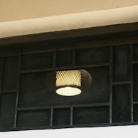 A patterned lamp shade with a glowing light bulb is visible through a grid-like wire mesh or frame. The setting appears to be indoors with a muted and soft lighting atmosphere.