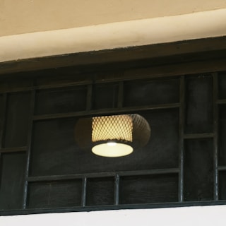 A patterned lamp shade with a glowing light bulb is visible through a grid-like wire mesh or frame. The setting appears to be indoors with a muted and soft lighting atmosphere.