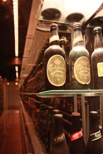 A close-up view of several beer bottles, primarily featuring Carlsberg Pilsner, arranged on a shelf. The bottles are illuminated by warm lighting, creating a cozy, inviting ambiance. The background shows rows of similar bottles, suggesting a display or storage area, possibly in a bar or a brewery.