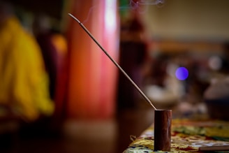 A single incense stick is placed in a wooden holder on a table. The incense is lit, with gentle smoke rising from the tip. The background is blurred, showcasing warm tones and hints of people and furniture, contributing to a calm and serene atmosphere.
