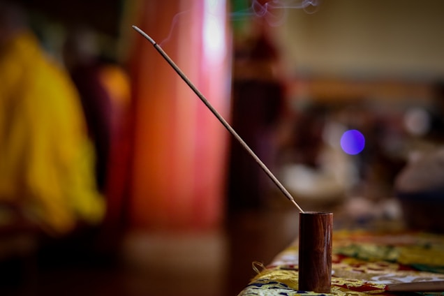 A single incense stick is placed in a wooden holder on a table. The incense is lit, with gentle smoke rising from the tip. The background is blurred, showcasing warm tones and hints of people and furniture, contributing to a calm and serene atmosphere.