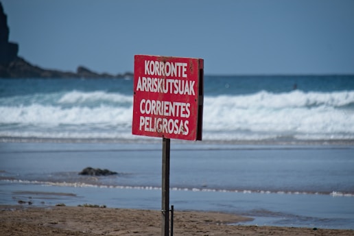 A red warning sign is prominently displayed on a beach, cautioning about dangerous currents. The ocean waves roll in the background, and a rocky outcrop is visible in the distance.