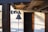 A rustic scene featuring a wooden beam supporting a decorative metal bell with three bird figures on top. The background includes a wooden door and a cream-colored wall illuminated by sunlight, with visible wooden beams on the ceiling. A small wall-mounted lamp is present near the door.