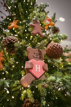 A festive Christmas tree adorned with warm white lights and wooden ornaments in the shapes of gingerbread people, pine cones, and a teddy bear holding a red heart with a letter 'H'. The branches are lush and green, providing a vibrant backdrop for the decorations.