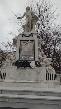 A large, ornate statue featuring a prominent central figure, possibly a historical or cultural figure, surrounded by an elaborate base with smaller sculptures of cherubic figures and musical motifs. The monument is situated in an outdoor setting, with bare trees visible in the background, suggesting a winter or late autumn scene.