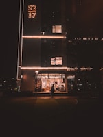A dimly lit urban street scene at night, featuring a building with illuminated signage reading 'S2 17'. Below, a well-lit bakery named 'Paris Gateaux' is visible with people entering and exiting. The scene is framed by street lamps and trees, creating a cozy urban atmosphere.
