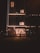 A dimly lit urban street scene at night, featuring a building with illuminated signage reading 'S2 17'. Below, a well-lit bakery named 'Paris Gateaux' is visible with people entering and exiting. The scene is framed by street lamps and trees, creating a cozy urban atmosphere.