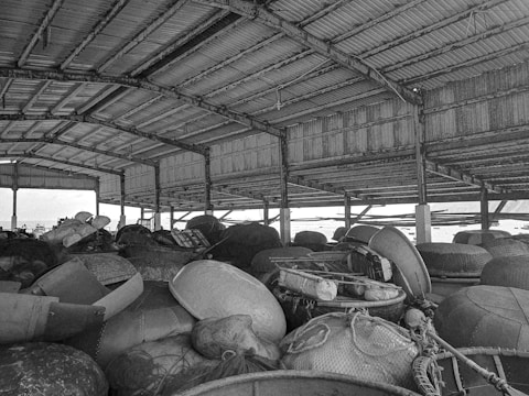 A storage area with a corrugated metal roof holding various round baskets and fishing equipment. The space is cluttered with numerous items arranged haphazardly. Some light filters through the gaps in the structure, indicating an outdoor environment beyond the enclosure.
