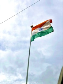 An Indian national flag is mounted on a tall flagpole, waving against a backdrop of a cloudy sky. The tricolor consists of saffron, white, and green horizontal stripes with a blue Ashoka Chakra in the center.