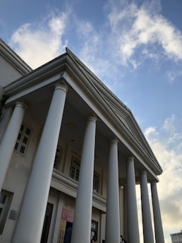A classical building with large, white, Greco-Roman columns and a pediment, set against a partly cloudy blue sky. There are windows and doors visible on the building's facade, and people can be seen near the entrance.