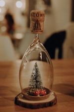 A glass ornament in the shape of an inverted wine glass contains a miniature artificial Christmas tree and small decorative mushrooms. It stands on a wooden base, with a cork stopper balanced on top. The scene is softly lit, with bokeh lights in the background creating a warm, cozy ambiance.
