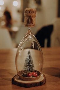 A glass ornament in the shape of an inverted wine glass contains a miniature artificial Christmas tree and small decorative mushrooms. It stands on a wooden base, with a cork stopper balanced on top. The scene is softly lit, with bokeh lights in the background creating a warm, cozy ambiance.