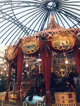A whimsical indoor carousel with ornately decorated panels and rich red drapery. The ceiling structure is a dome of dark metal beams, adding to the vintage atmosphere. Bright lights hang from the structure, reflecting off the bronze and gold accents. Beneath the carousel, a small blue vintage car is parked, surrounded by a low fence and foliage. Baskets and various decorative elements are incorporated into the design.