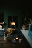 A cozy living room featuring a lit wood stove emanating a warm glow. The space includes a dark rug, a small coffee table with a candle and mug, and a wicker basket on the floor. There is also a chunky knit blanket in mustard yellow color on the rug. The room has a dark, moody ambiance with subtle holiday decor, like a small decorated Christmas tree.
