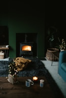 A cozy living room featuring a lit wood stove emanating a warm glow. The space includes a dark rug, a small coffee table with a candle and mug, and a wicker basket on the floor. There is also a chunky knit blanket in mustard yellow color on the rug. The room has a dark, moody ambiance with subtle holiday decor, like a small decorated Christmas tree.