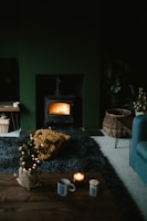 A cozy living room featuring a lit wood stove emanating a warm glow. The space includes a dark rug, a small coffee table with a candle and mug, and a wicker basket on the floor. There is also a chunky knit blanket in mustard yellow color on the rug. The room has a dark, moody ambiance with subtle holiday decor, like a small decorated Christmas tree.