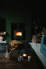 A cozy living room featuring a lit wood stove emanating a warm glow. The space includes a dark rug, a small coffee table with a candle and mug, and a wicker basket on the floor. There is also a chunky knit blanket in mustard yellow color on the rug. The room has a dark, moody ambiance with subtle holiday decor, like a small decorated Christmas tree.