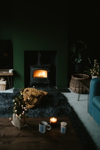 A cozy living room featuring a lit wood stove emanating a warm glow. The space includes a dark rug, a small coffee table with a candle and mug, and a wicker basket on the floor. There is also a chunky knit blanket in mustard yellow color on the rug. The room has a dark, moody ambiance with subtle holiday decor, like a small decorated Christmas tree.