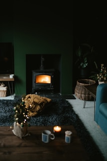 A cozy living room featuring a lit wood stove emanating a warm glow. The space includes a dark rug, a small coffee table with a candle and mug, and a wicker basket on the floor. There is also a chunky knit blanket in mustard yellow color on the rug. The room has a dark, moody ambiance with subtle holiday decor, like a small decorated Christmas tree.
