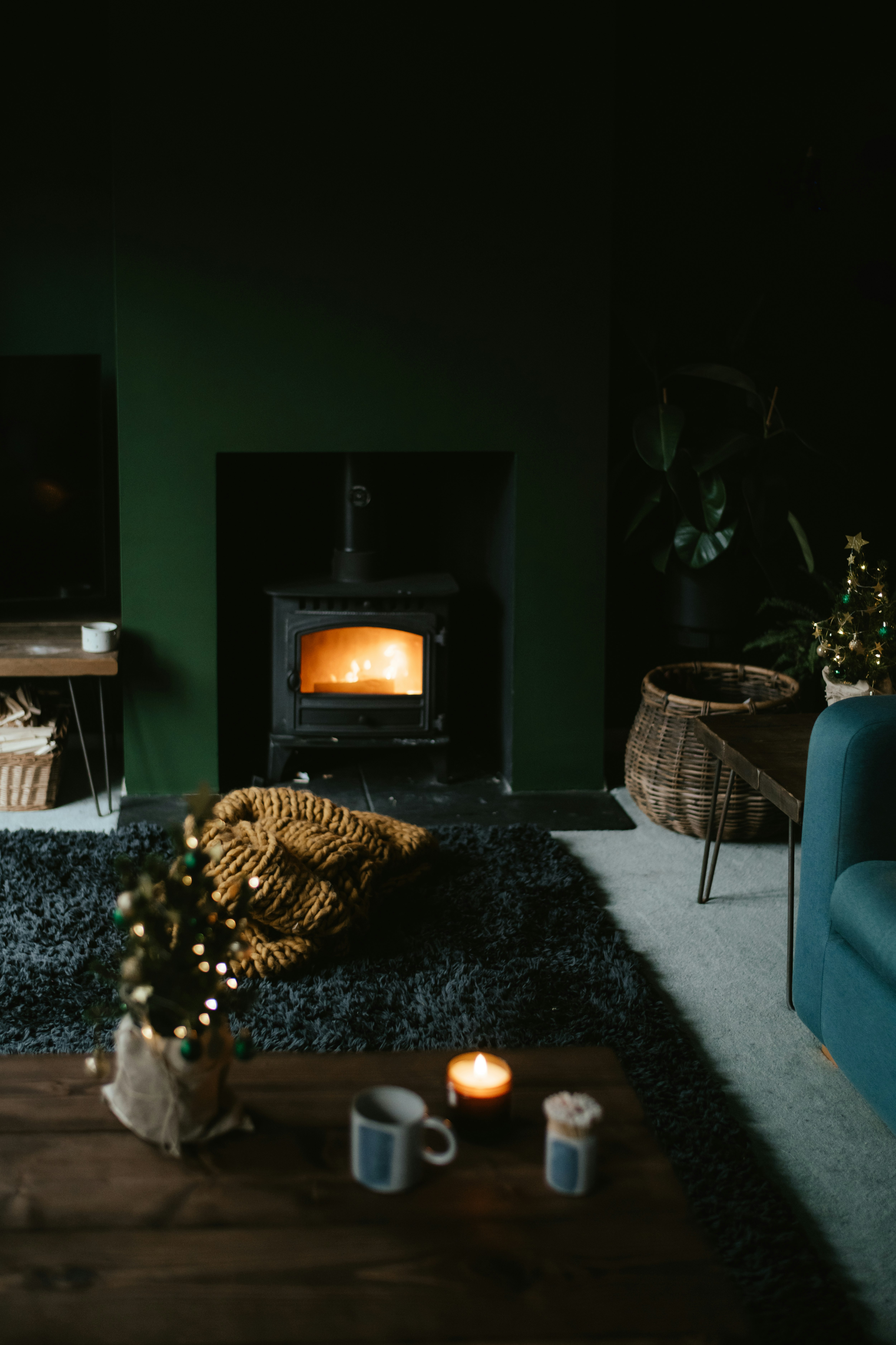 A cozy living room featuring a lit wood stove emanating a warm glow. The space includes a dark rug, a small coffee table with a candle and mug, and a wicker basket on the floor. There is also a chunky knit blanket in mustard yellow color on the rug. The room has a dark, moody ambiance with subtle holiday decor, like a small decorated Christmas tree.