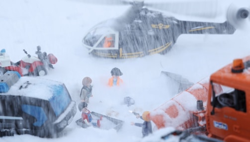 A snowy scene with toy figures involved in a rescue operation. A helicopter hovers above, while people dressed in rescue gear are engaged in activities on the ground. There are toy vehicles like an ATV and a rescue truck covered in snow. The blizzard creates a sense of urgency and action.