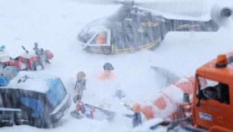 A snowy scene with toy figures involved in a rescue operation. A helicopter hovers above, while people dressed in rescue gear are engaged in activities on the ground. There are toy vehicles like an ATV and a rescue truck covered in snow. The blizzard creates a sense of urgency and action.