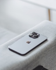 A modern smartphone with a sleek design and three camera lenses on the back is placed on a light gray fabric surface. The device features a recognizable apple logo.