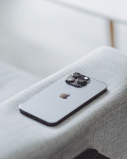 A modern smartphone with a sleek design and three camera lenses on the back is placed on a light gray fabric surface. The device features a recognizable apple logo.