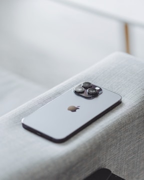 A modern smartphone with a sleek design and three camera lenses on the back is placed on a light gray fabric surface. The device features a recognizable apple logo.