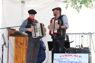 Two men in casual attire play accordions in an indoor setting. Both are wearing vests and flat caps. One has a red accordion, and the other has a beige one. Various musical equipment and microphones are present, and a sign reading 'Tyrol'Alp' is visible.