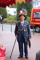 An elderly man is dressed in a decorated navy blue suit covered with numerous pins and badges. He wears a vibrant, multicolored hat adorned with flowers and various decorations, including a small French flag. The man holds a walking stick in one hand and an umbrella in the other. In the background, there is a bright red emergency vehicle and some lush greenery.