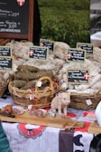 An outdoor market stall displaying baskets of cured sausages arranged neatly, with labels in French indicating the types of products. A small decorative pig figurine is placed on a wooden board along with sliced pieces of sausage. A rustic tablecloth with traditional designs covers the table, creating a homely, country market feel. A chalkboard sign in the background provides pricing information.