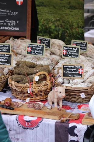 An outdoor market stall displaying baskets of cured sausages arranged neatly, with labels in French indicating the types of products. A small decorative pig figurine is placed on a wooden board along with sliced pieces of sausage. A rustic tablecloth with traditional designs covers the table, creating a homely, country market feel. A chalkboard sign in the background provides pricing information.
