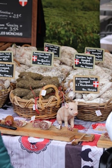An outdoor market stall displaying baskets of cured sausages arranged neatly, with labels in French indicating the types of products. A small decorative pig figurine is placed on a wooden board along with sliced pieces of sausage. A rustic tablecloth with traditional designs covers the table, creating a homely, country market feel. A chalkboard sign in the background provides pricing information.