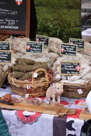 An outdoor market stall displaying baskets of cured sausages arranged neatly, with labels in French indicating the types of products. A small decorative pig figurine is placed on a wooden board along with sliced pieces of sausage. A rustic tablecloth with traditional designs covers the table, creating a homely, country market feel. A chalkboard sign in the background provides pricing information.