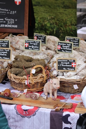 An outdoor market stall displaying baskets of cured sausages arranged neatly, with labels in French indicating the types of products. A small decorative pig figurine is placed on a wooden board along with sliced pieces of sausage. A rustic tablecloth with traditional designs covers the table, creating a homely, country market feel. A chalkboard sign in the background provides pricing information.