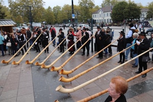A group of people is standing in an outdoor area holding long wooden alpine horns. They appear to be dressed in traditional attire, and many have hats. There is a crowd of spectators observing the scene. The setting is a paved public square, with trees and buildings visible in the background.