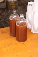 Two transparent bottles filled with a brown liquid are placed on a wooden surface. Next to them, there is a stack of white disposable cups. The background includes a blurry view of paving stones and cardboard boxes.