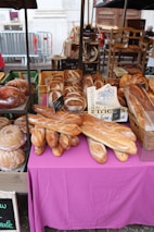 A market stall showcasing various types of freshly baked bread. There are several baguettes placed on a pink tablecloth, with a variety of loaves, including round and rectangular shapes. Labels with prices are visible on some of the bread. The background includes wooden shelves and boxes, with more bread displayed.
