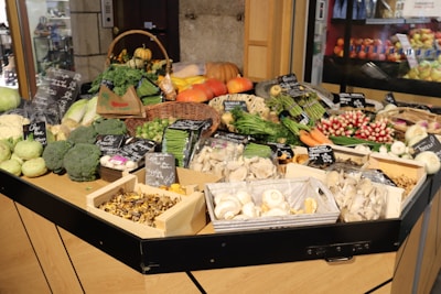 A market stall displays a variety of fresh vegetables including broccoli, asparagus, radishes, carrots, mushrooms, and leafy greens. Several black signs with handwritten labels are visible, indicating the names of the produce. Behind the vegetables, there are pumpkins and other squash varieties. The setup is indoors, with a refrigerated fruit section visible in the background.