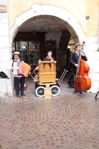Three street musicians perform under an archway. One plays an accordion, the second plays a barrel organ, and the third plays an upright bass. They are dressed in vintage clothing, and the background includes a cobblestone street and a shop entrance.