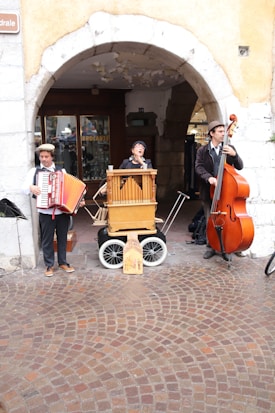 Three street musicians perform under an archway. One plays an accordion, the second plays a barrel organ, and the third plays an upright bass. They are dressed in vintage clothing, and the background includes a cobblestone street and a shop entrance.