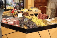 A market stall displaying a variety of fresh fruits including red and green grapes, blueberries, raspberries, strawberries, and apples. The fruits are arranged neatly in baskets and trays with small blackboards labeling some of the items. The setting is indoors with a wooden door and stone wall in the background.