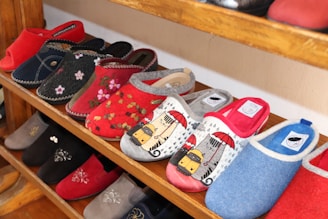A variety of colorful slippers are displayed on wooden shelves. The slippers feature different designs, including floral patterns, solid colors, and graphic prints such as animals under umbrellas. They are arranged neatly in rows, showcasing a range of styles and colors.