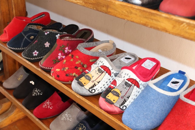 A variety of colorful slippers are displayed on wooden shelves. The slippers feature different designs, including floral patterns, solid colors, and graphic prints such as animals under umbrellas. They are arranged neatly in rows, showcasing a range of styles and colors.