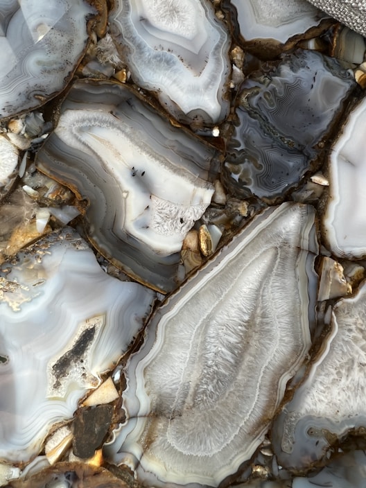 A close-up view of polished agate stones with intricate patterns and layers. The stones display a variety of gray, white, and brown tones with some areas appearing translucent. The natural formations create a mesmerizing and artistic appearance.