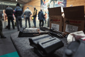 A table is covered with ammunition, including magazines and various bullets. Boxes labeled 'Wolf Military Classic' are visible, suggesting a setting related to firearms. In the background, several people are standing and appear to be engaged in discussion or activity within what looks like an indoor range or training facility. The setting suggests a focus on firearms training or practice.