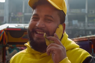 A man dressed in a yellow hoodie and cap is smiling and holding a banana to his ear, mimicking a phone. He is standing in an outdoor setting with colorful decorations and blurred buildings in the background.